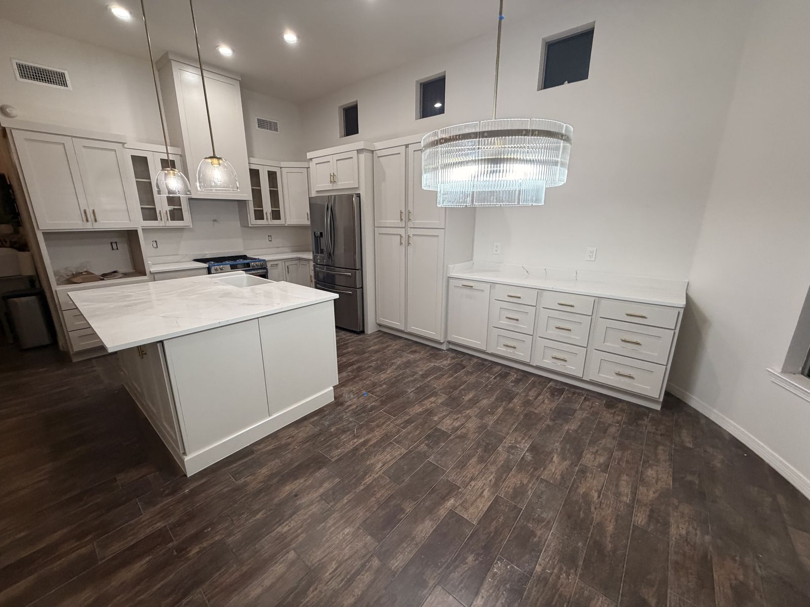 Large white kitchen with island, crystal chandelier and dark wood-look floors