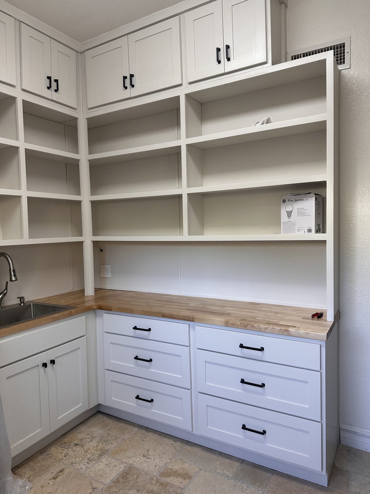 Corner of white shaker drawers with black pulls under a butcher-block countertop, with open shelving above